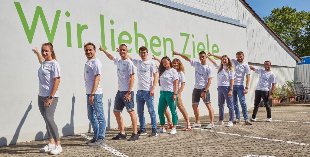 Alpensped Mitarbeitende zeigen auf den Schriftzug „Wir lieben Ziele“
